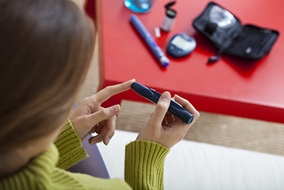 Photo taken from slightly above and behind a person wearing a green turtleneck with long sleeves. They are holding a lancing device to their finger. The table in front of them has several additional tools diabetics use, including a glucometer, and an open carrying case.