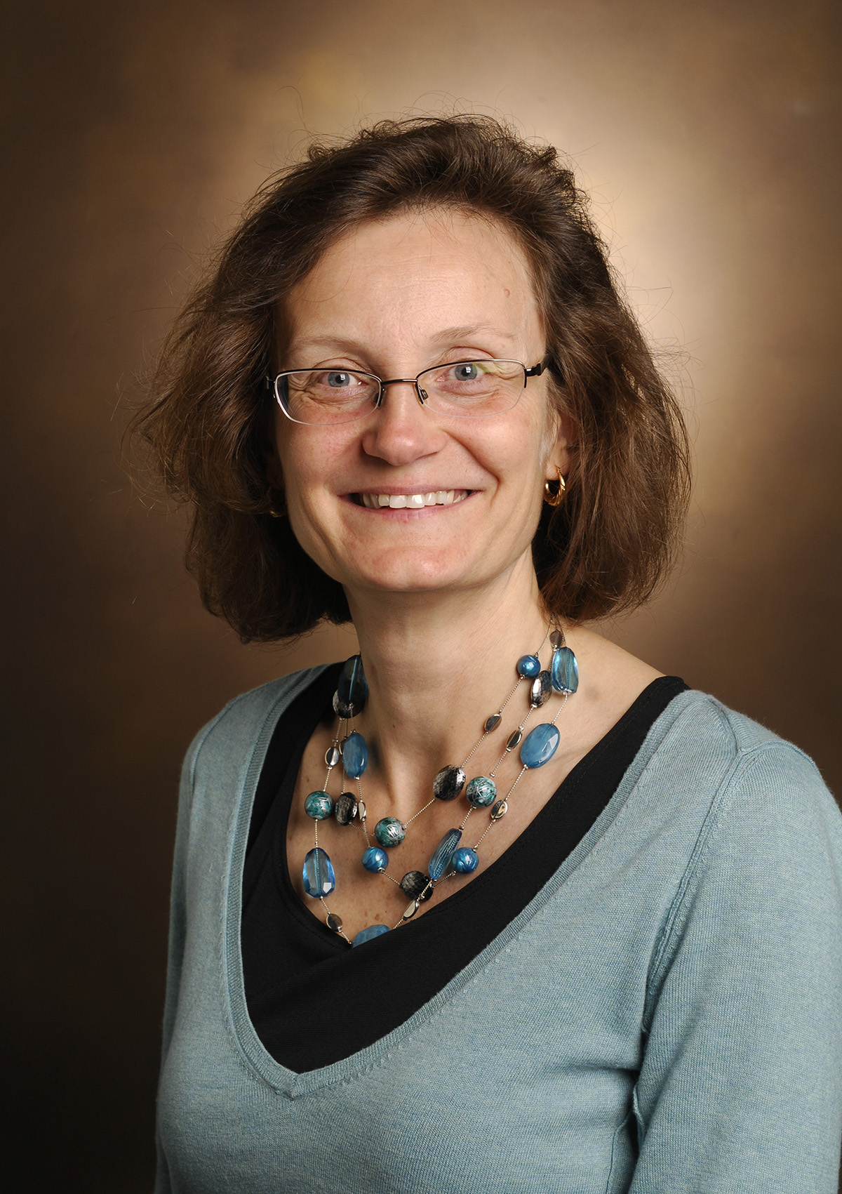 Photo of Ambra Pozzi with eye-glasses. She is wearing a light blue sweater over a black top and is also wearing a necklace made of blue and black stones.