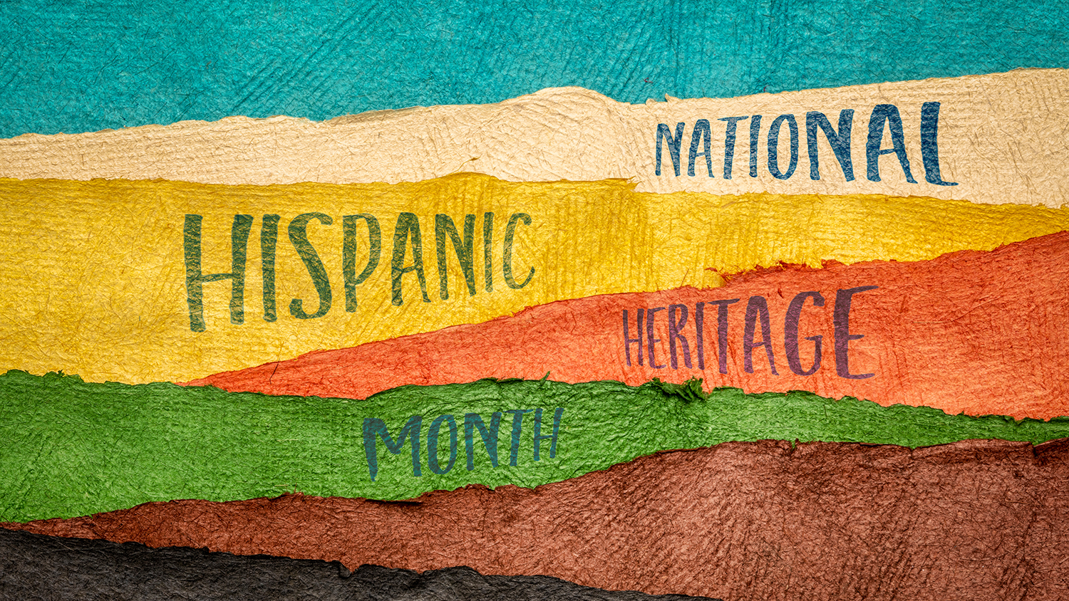 Handwriting that says "National Hispanic Heritage Month" on Huun paper handmade in Mexico. Strips of colorful paper are overlaid horizontally. From top to bottom, the strips are teal, tan, yellow, reddish orange, green, brown, dark brown. The words are written on the central layers.