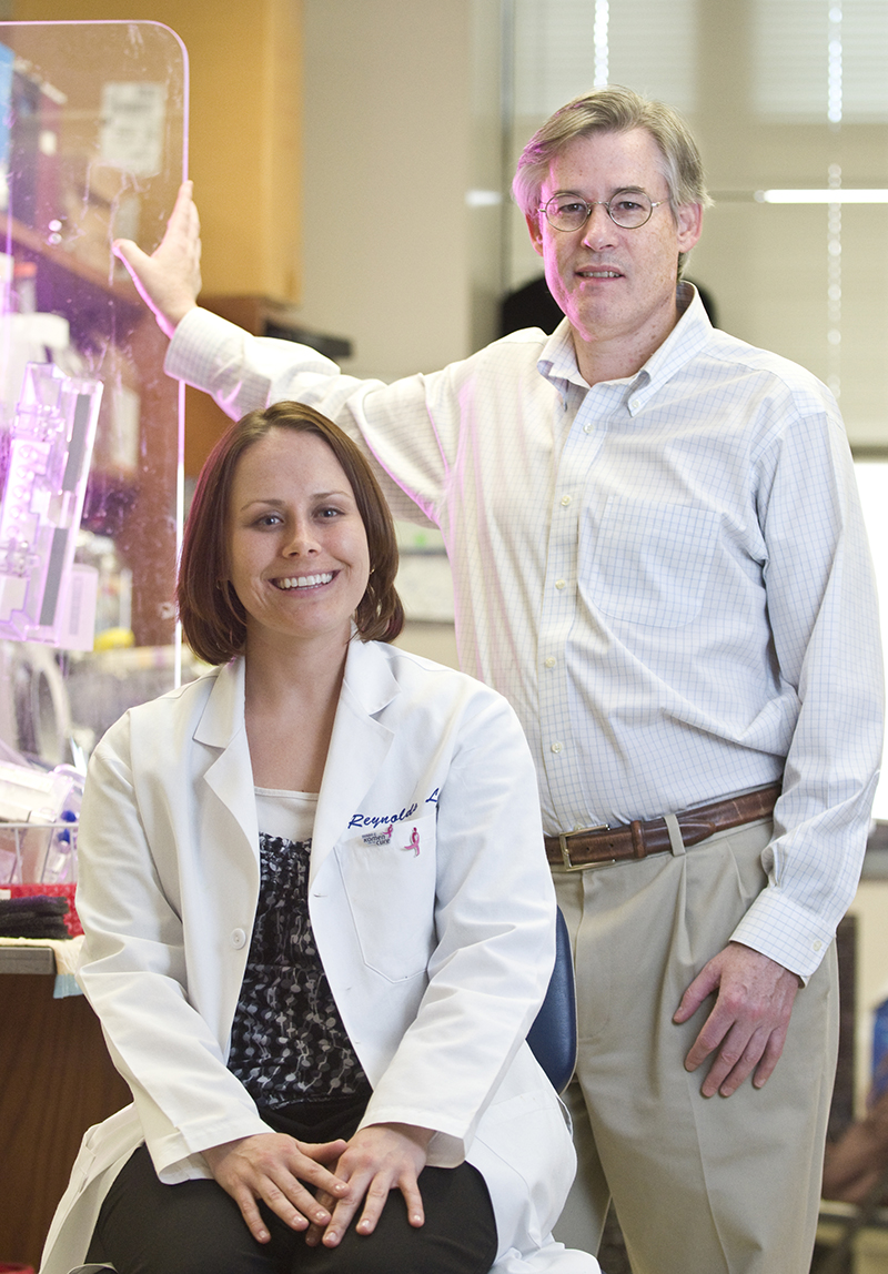 Sarah Kurley and Al Reynolds photographed in Reynolds' lab. Kurley is sitting down and Reynolds is standing next to her and slightly behind. Kurley is wearing a lab coat with two breast cancer awareness pins, and Reynolds is wearning slacks and a light-colored collared shirt.