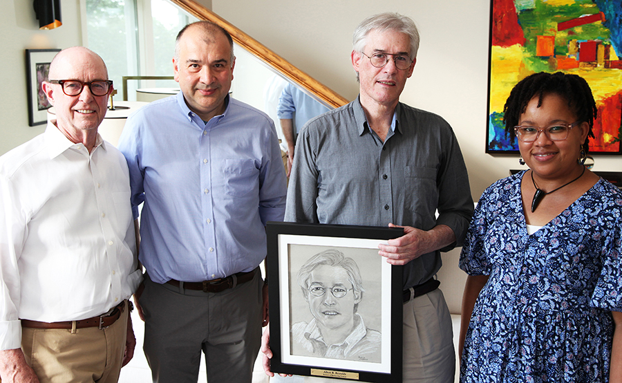 Four people standing next to one another. From left to right: Larry Marnett, Ege Kavalali, Al Reynolds, and Nadine Shillingford. Reynolds is holding a framed pencil portrait of himself.