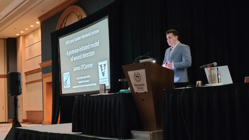 James O'Connor standing at a podium with the Sheraton logo on it on an elevated platform. A screen on his right has the title slide of his presentation.