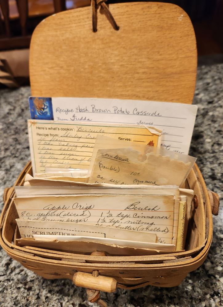 A basket-style recipe box with a lid and many handwritten recipes sticking out from inside it. The box is sitting on a granite countertop.