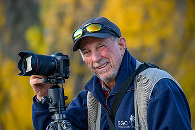 Guengerich smiling while holding a camera sitting on a tripod.