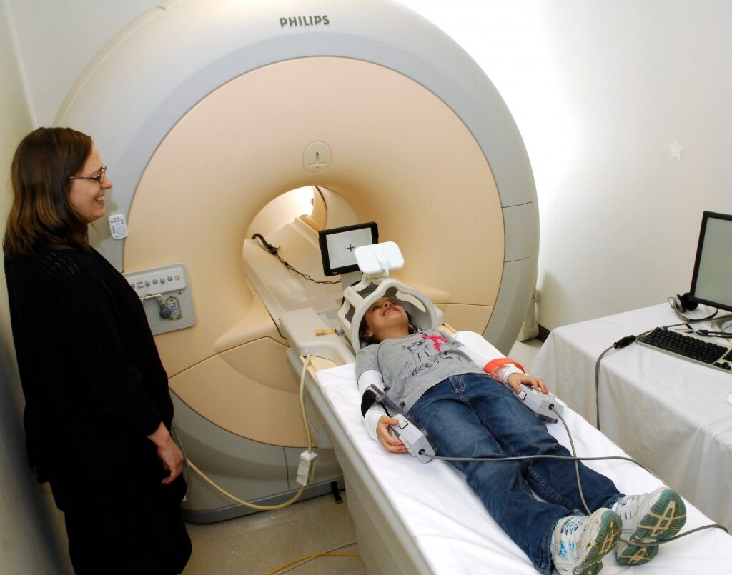 A smiling child is lying on a mock MRI machine with an adult looking on, smiling.