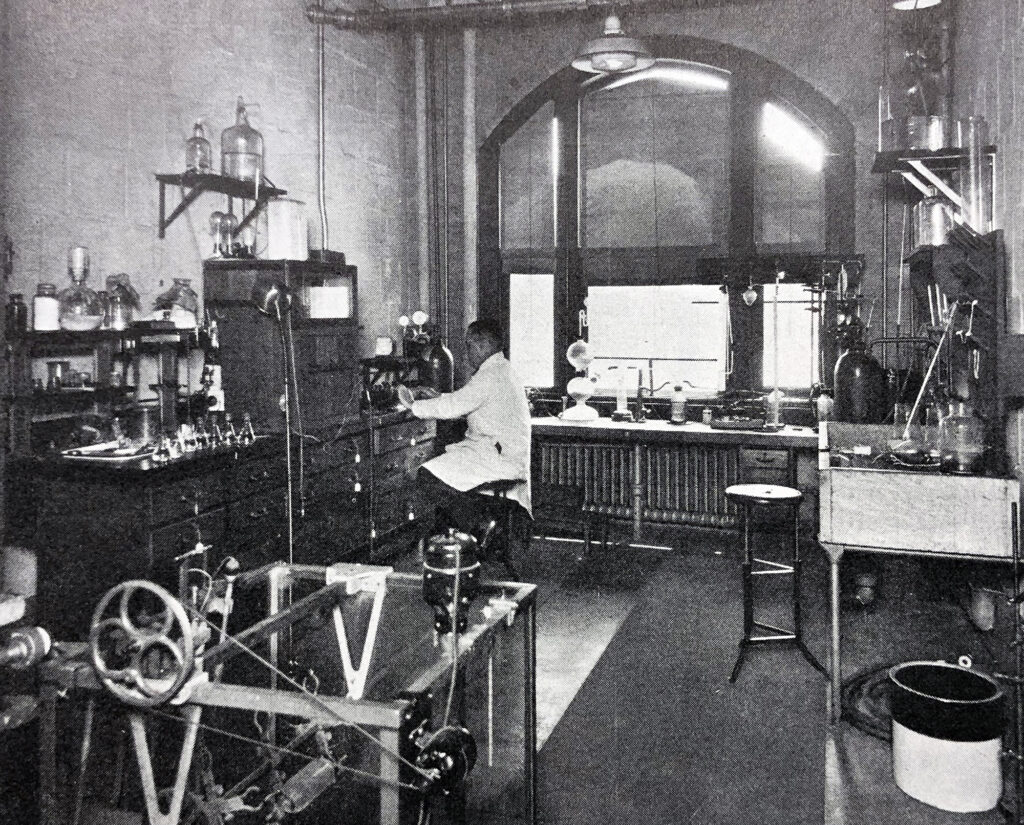 A black and white photo of a researcher in a lab. There is glass equipment throughout and some sort of machinery in the foreground. 