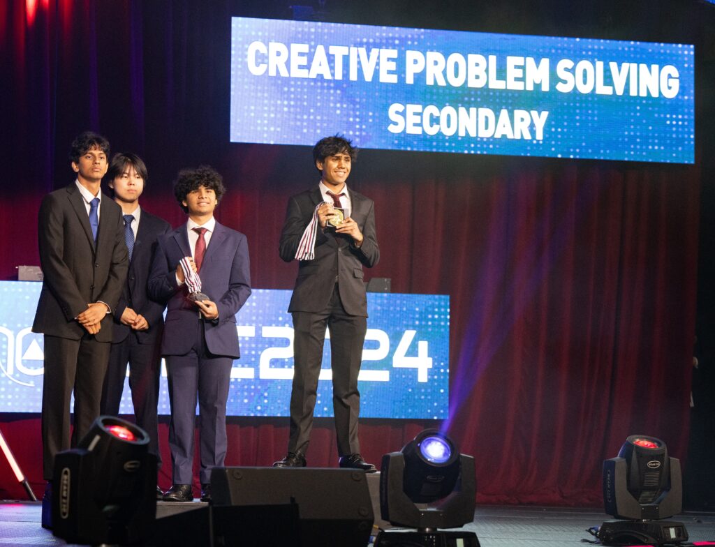 Teebro Paul and two teammates, left, stand on the second-place step of a podium at the Creative Problem Solving award ceremony. Paul and the first-place finish are holding medals in their hands. All students are in suits, reflecting the formality of the event.