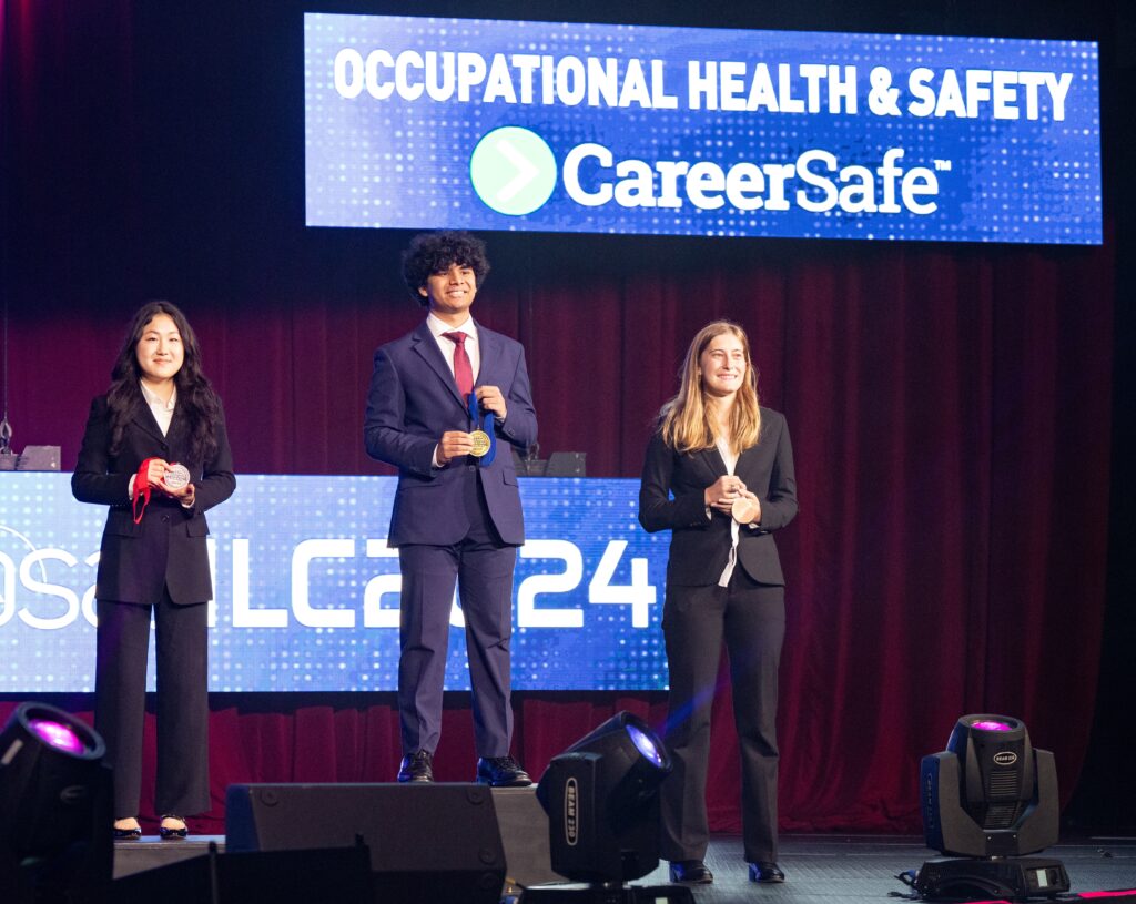 Teebro Paul, center, stands on the first-place step of a podium. He is flanked by the two students who won second and third place. All students are in suits, reflecting the formality of the event.