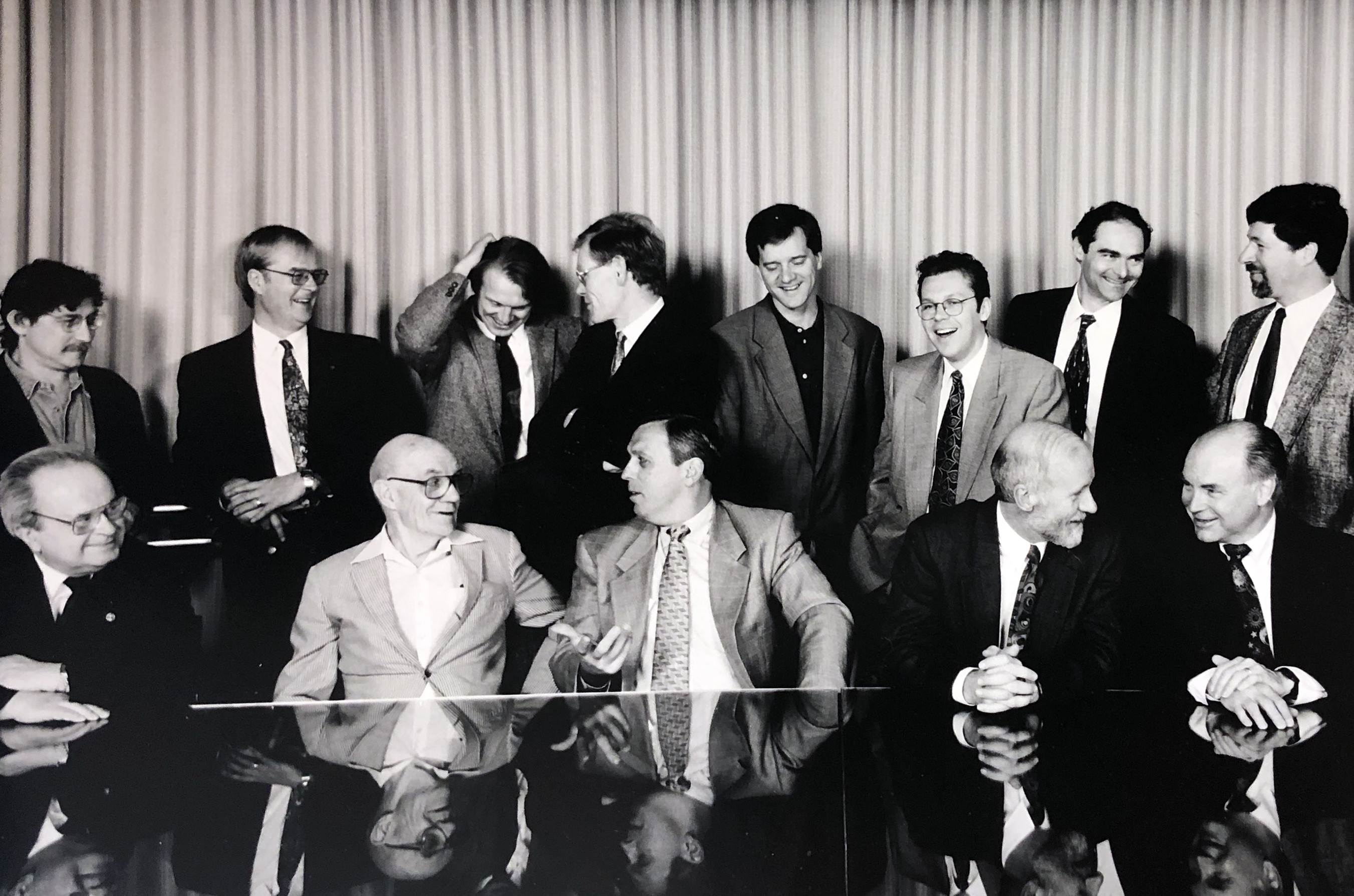 Black and white photograph of men sitting at table. Other men are standing behind them.