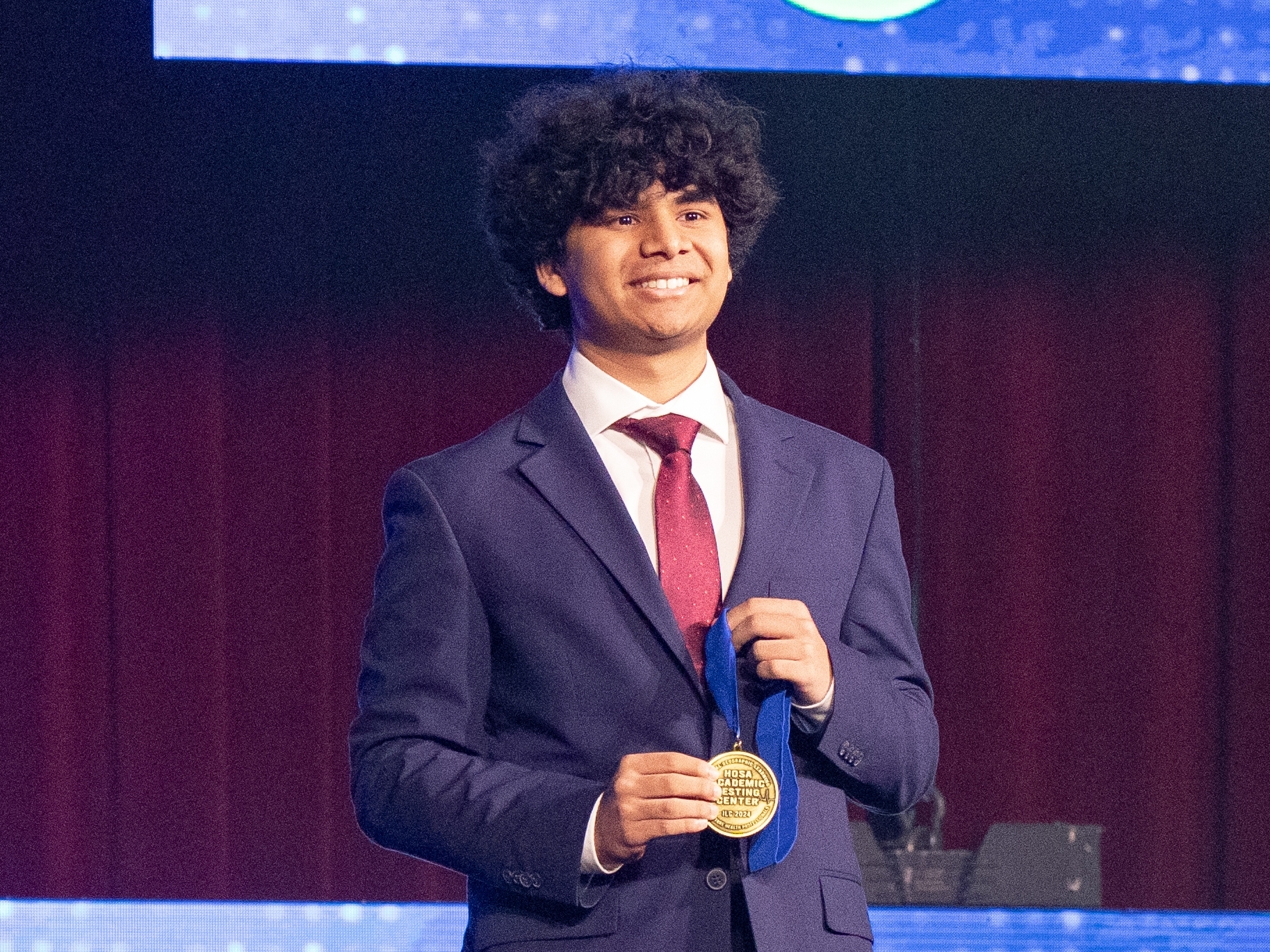 Teebro Paul, in a suit, holding a gold medal and smiling.