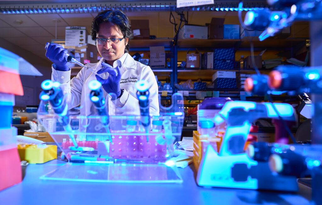 Mirazul Islam wearing gloves and a lab coat pipetting something in a microcentrifuge tube. The shot is taken from the other side of the bench so you can see lab equipment all around Islam. The photo has a blue hue throughout.