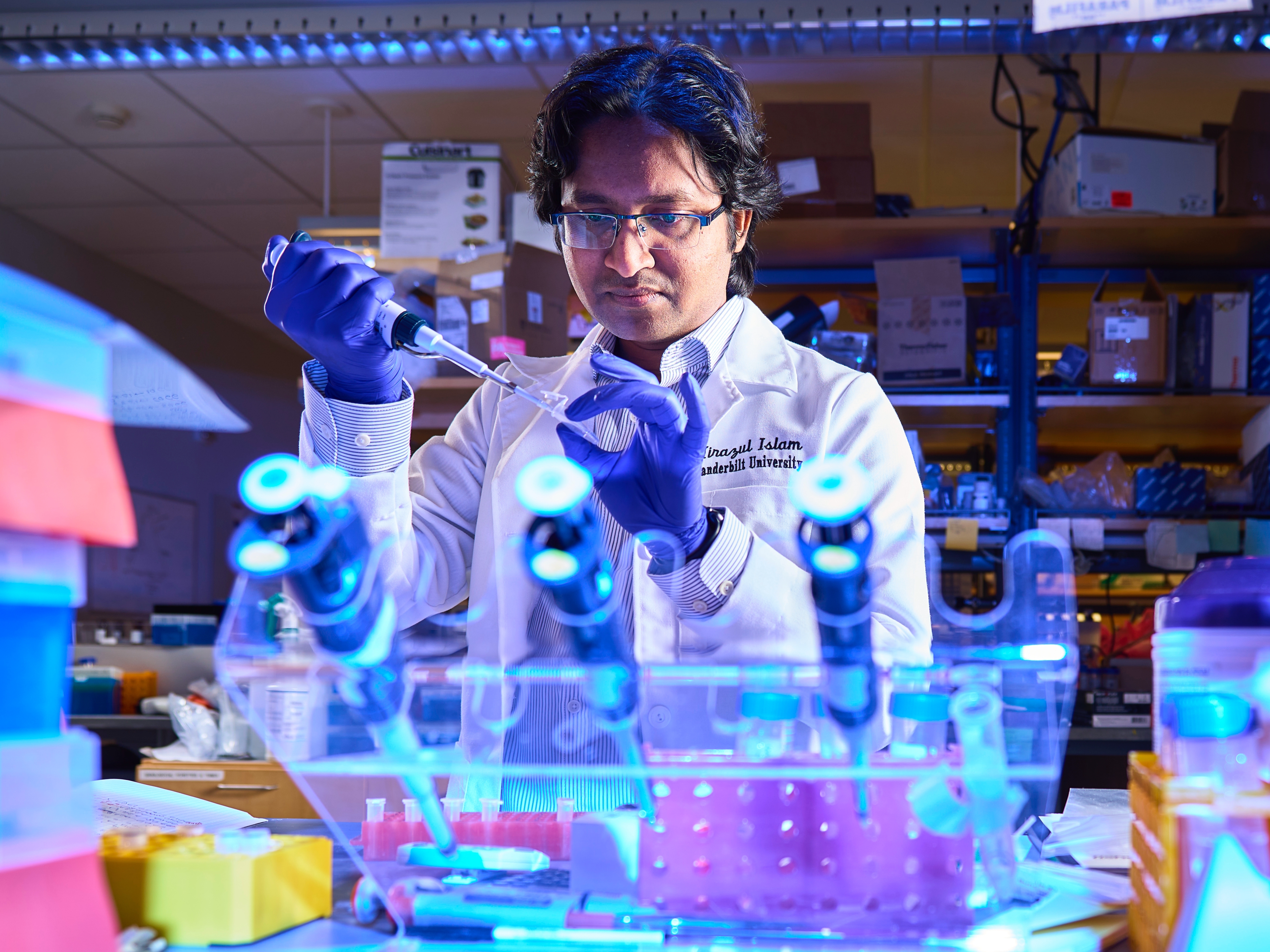 Mirazul Islam wearing gloves and a lab coat pipetting something in a microcentrifuge tube. The shot is taken from the other side of the bench so you can see lab equipment all around Islam. The photo has a blue hue throughout.