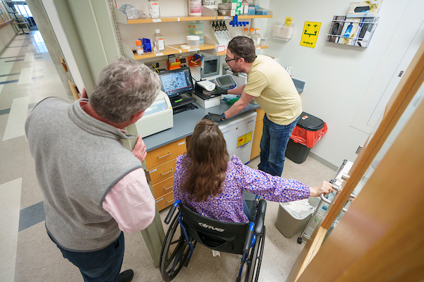 Three people looking at a computer monitor. The person in the middle is in a wheelchair