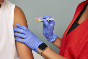 Closeup of doctor's hands, vaccine, and arm. The doctor is wearing blue nitrile groves and a smart watch. The syringe is uncapped - the doctor is holding it as if about to give the patient the shot.