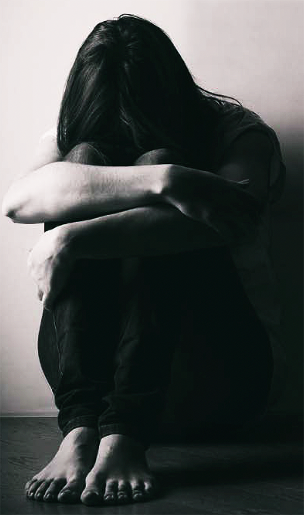 A black and white photograph of a person with long hair sitting on the floor, hugging their knees, and holding their head on their knees. The image is a visualization of someone who is depressed.