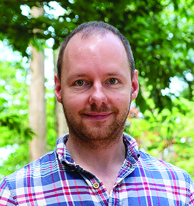 Headshot of Alexander Thiemicke. He's wearing a plaid, collared shirt and is posing in front of some trees/greenery.