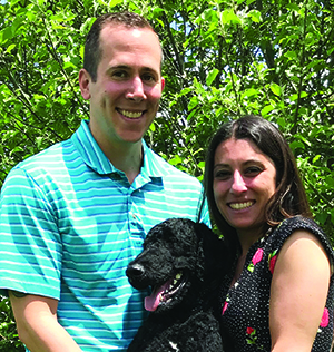 Photo of Dan Quimby in horizontal striped shirt (left) and his wife (right) holding a large black dog (center).