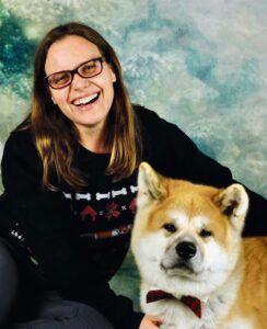 Armelle Le Guelte, wearing a black sweater with white bones and additional red printed items on it (looks to be an "ugly" holiday sweater), next to her dog, a fluffy white and gold thing with a red bowtie.