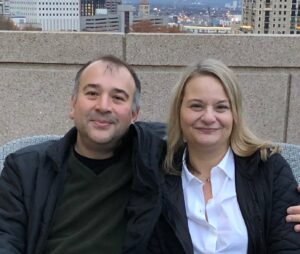 Ege Kavalali (left) and Lisa Monteggia (right) sitting on a gray wicker chair on a balcony. Over the edge of the stone balcony you can see Nashville. Kavalali is wearing a dark sweater and a black jacket and Monteggia is wearing a white blouse and a black jacket.