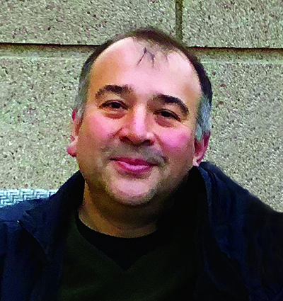 Headshot of Ege Kavalali. He is wearing a black coat. He sits in front of a concrete wall.