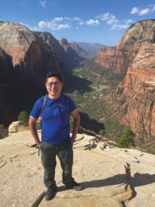 Geoffrey Li standing atop a rock surface with a canyon and a green valley in the background. Li is wearing hiking pants, a blue t-shirt, and a backpack.
