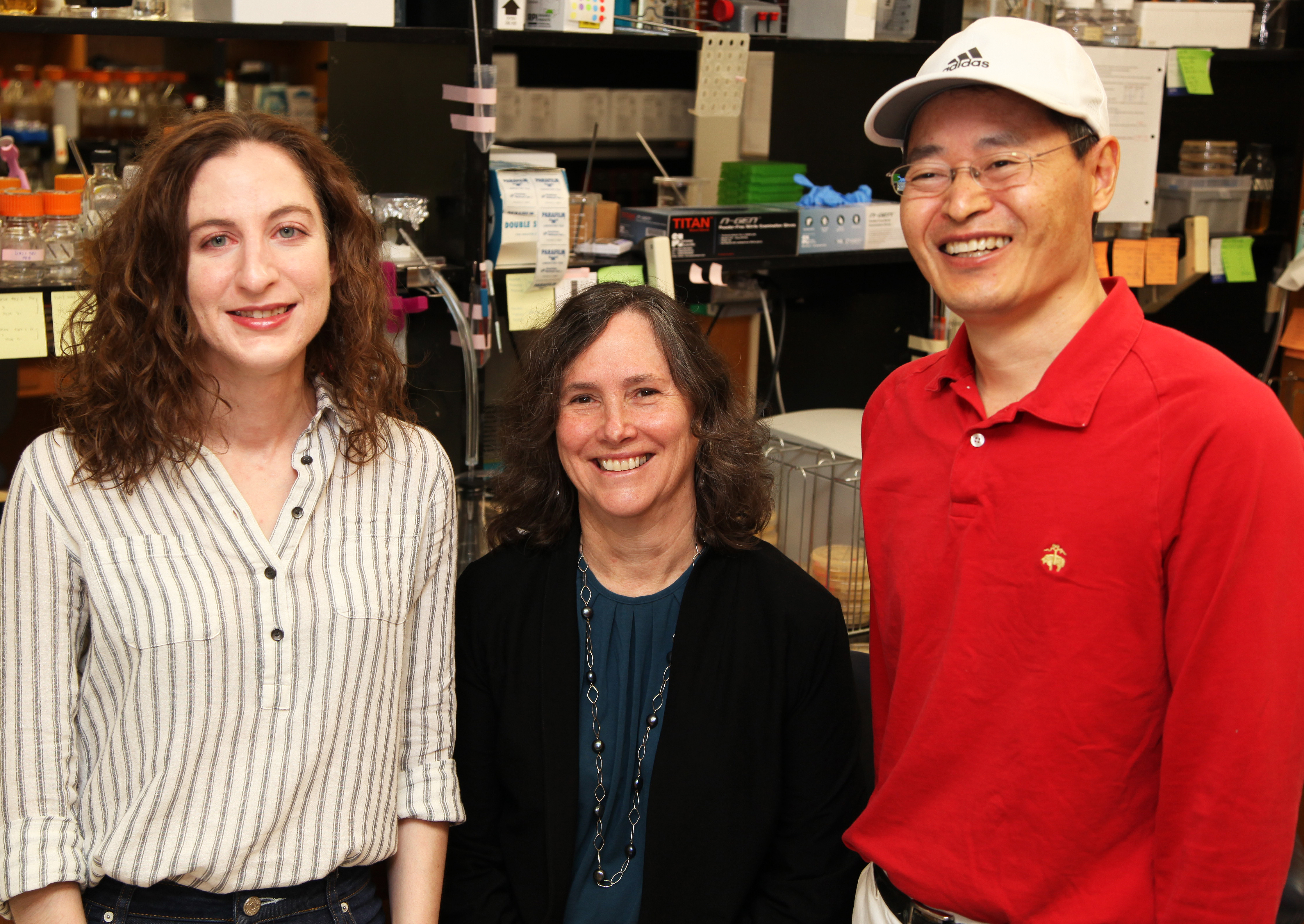 Photo of Sierra Cullati, Kathy Gould, and Jun-Song Chen in the Gould lab. Gould is sitting down, flanked by Cullati on the left and Chen on the right. Cullati is wearing a striped blouse, Gould a turquoise blouse and a black cardigan, and Chen a red, long-sleeved shirt and a baseball cap.