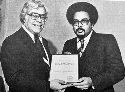 Black and white photo of Gilbert Ortiz (left) presenting an award to Michael Fant (right).