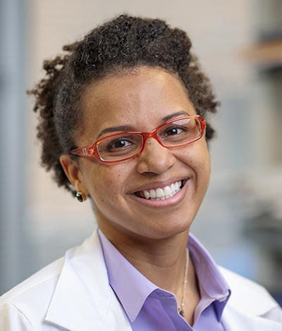 Headshot of Assistant professor Rita Brookheart, PhD wearing red-rimmed eye-glasses and a white lab coat over a lilac-colored shirt.