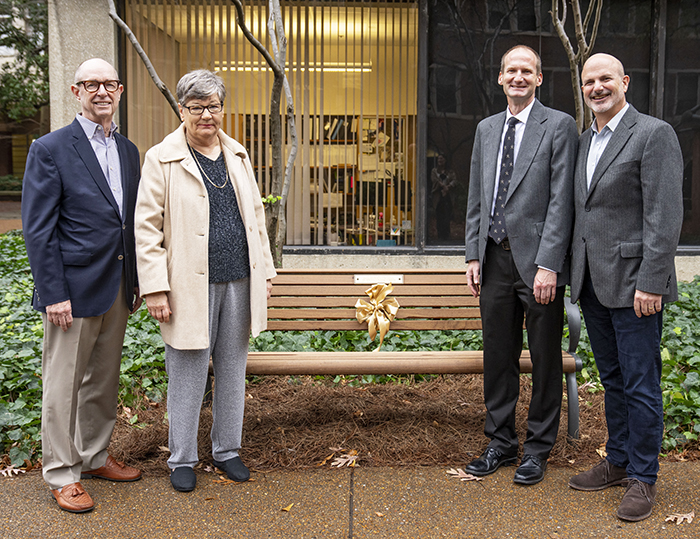 Photo of (L-R) Larry Marnett wearing a blue blazer, Jan Jordan wearing a cream-colored coat, David Cortez wearing a gray blazer, and John York wearing a gray blazer. They are standing in front of a bench which has a gold ribbon tied to its middle. 