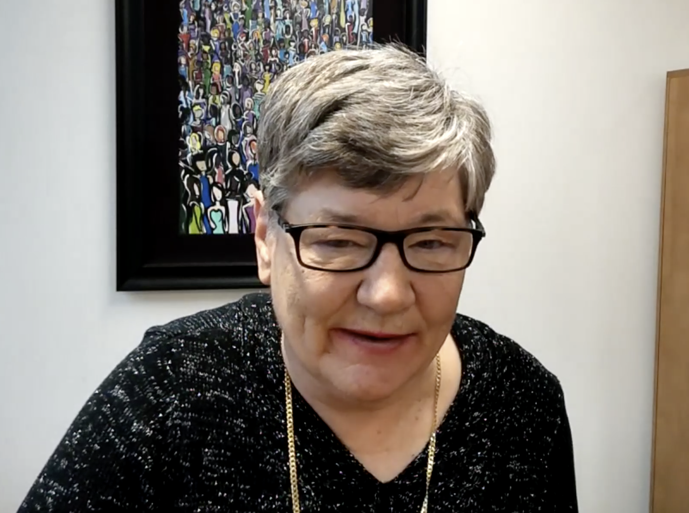 Photo of Jan Jordan giving a Zoom presentation. She is earring a black-and-white-flecked top and a gold necklace. Behind her is a painting of a crowd of people.