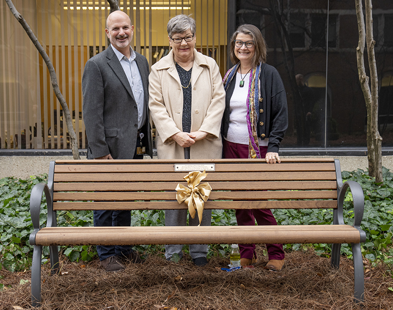 Photo of bench with a gold ribbon tide in the middle. Behind it stand John York wearing a gray blazer, Jan Jordan wearing a cream colored coat, and Sally York wearing a dark sweater and multi-colored scarf.