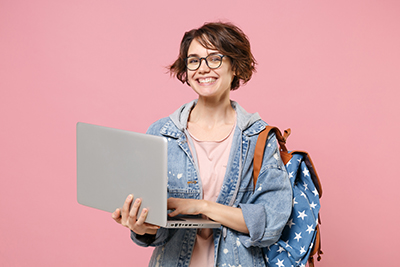 Cheerful young woman student in denim clothes glasses backpack isolated on pastel pink background. Education in high school university college concept. Mock up copy space. Work on laptop pc computer