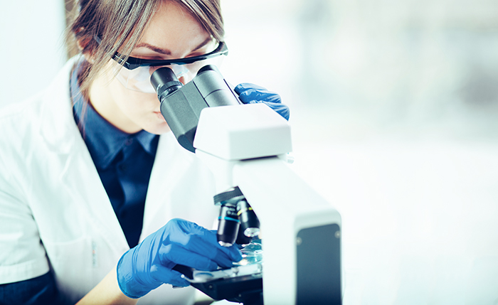 A scientist looks intently through a microscope. She has dark hair and is wearing a lab coat, gloves, and safety glasses.
