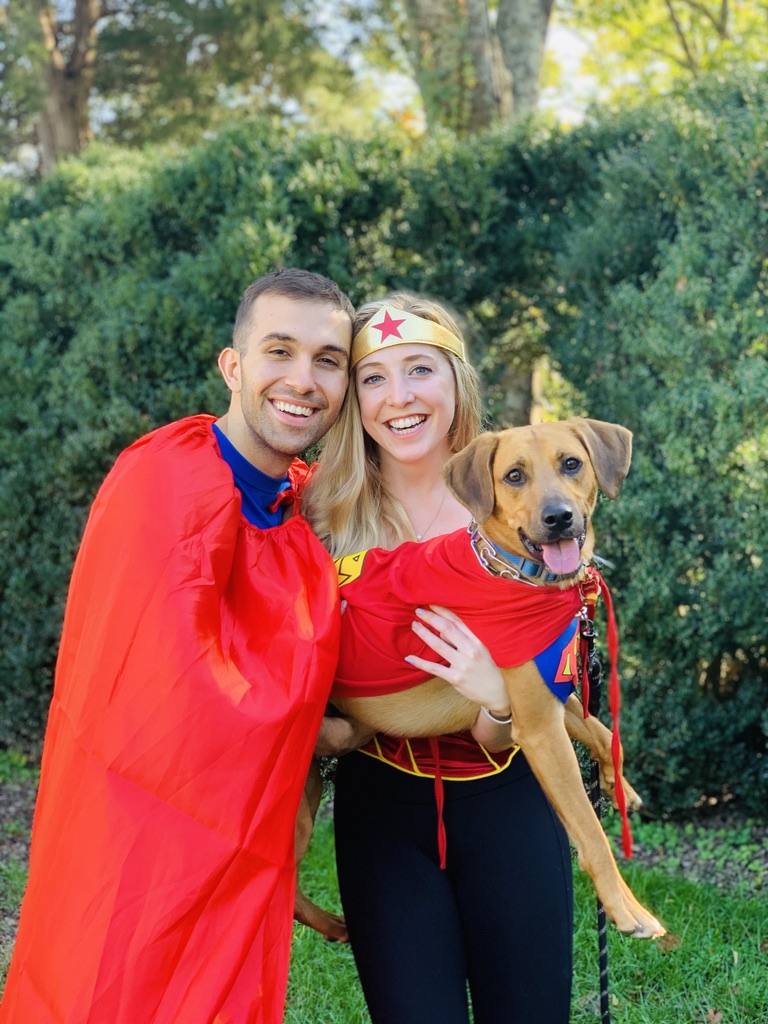 A man and a woman pose with a dog wearing a red cape. The three subjects are dressed as superheroes for Halloween.