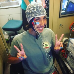A woman holds up two fingers on each hand while wearing an EEG cap