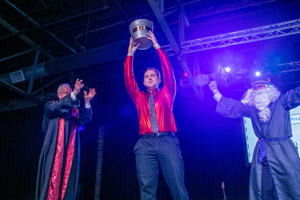 A man lifts a bucket on stage near to two costumed people
