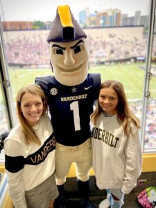 Two women stand with a Commodore mascot in a skybox at a football game