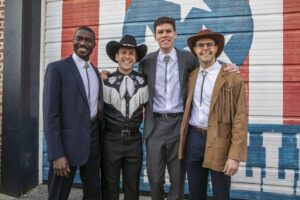 Four men in bolo ties and suits pose in front of a mural