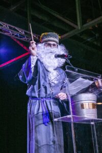 A man in a fake beard and robe holds a fake wand as he speaks into a microphone