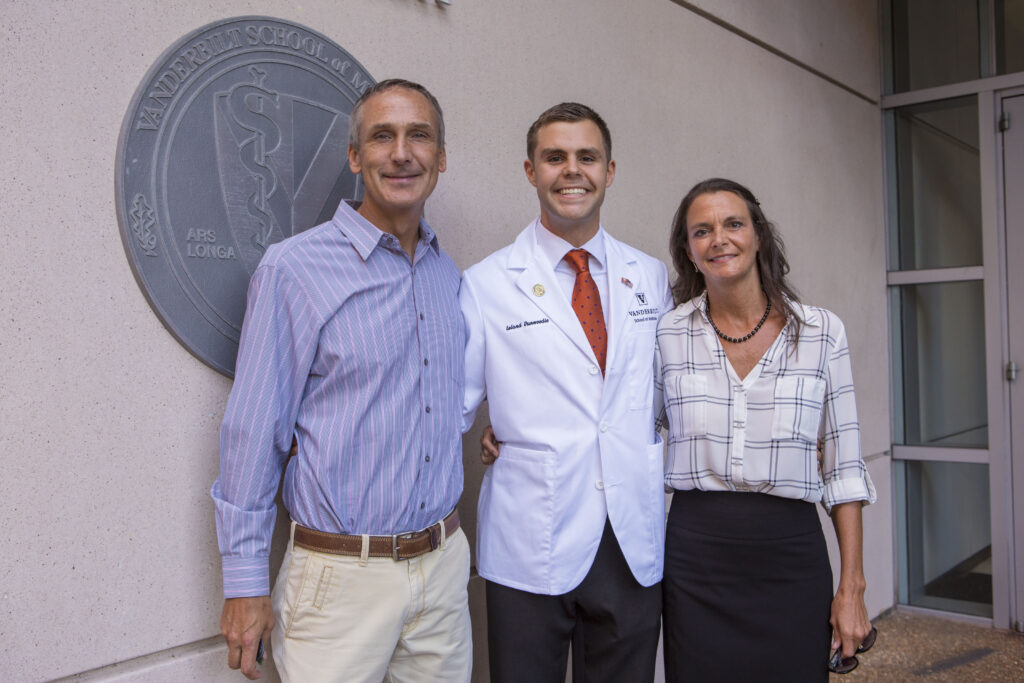 Three people pose outside a medical school building. A school seal hangs on the wall behind them.