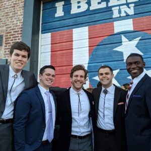 Five men in bolo ties and suits pose in front of a mural