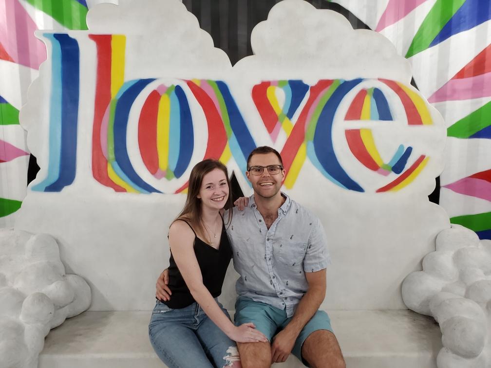 A man and woman sit on a white couch. A multicolored sign behind them says "love"