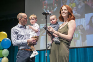 A man and a woman both hold baby boys while standing on a stage.