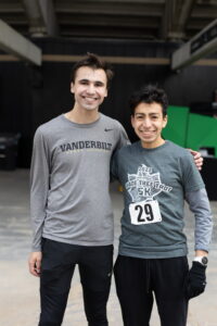 Two men in running clothes pose for a photo. One man wears a race bib.