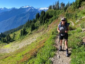 A woman with hiking poles stands on a green trail with mountains in the background.