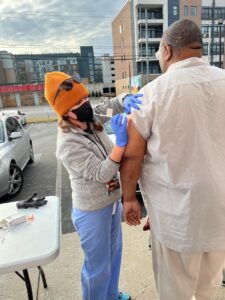 A woman in scrubs and a sweater gives a vaccine to a person wearing a white shirt