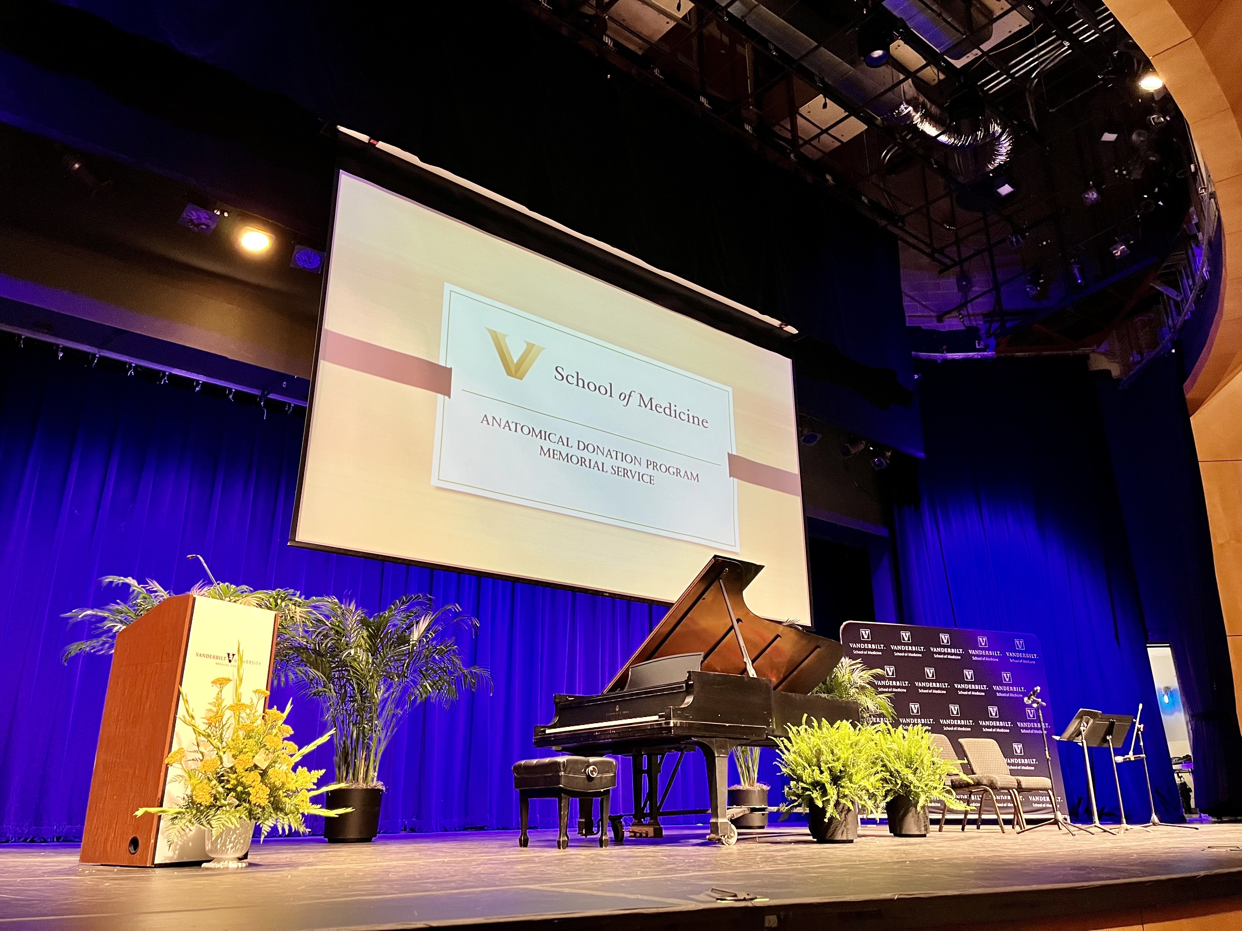 A piano sits on a stage with a lectern and chairs in front of a memorial service screen