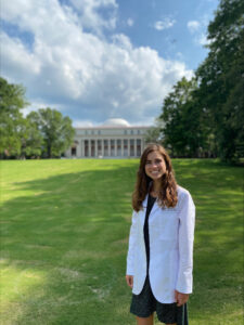 A woman in a white coat stands outside in front of an academic building