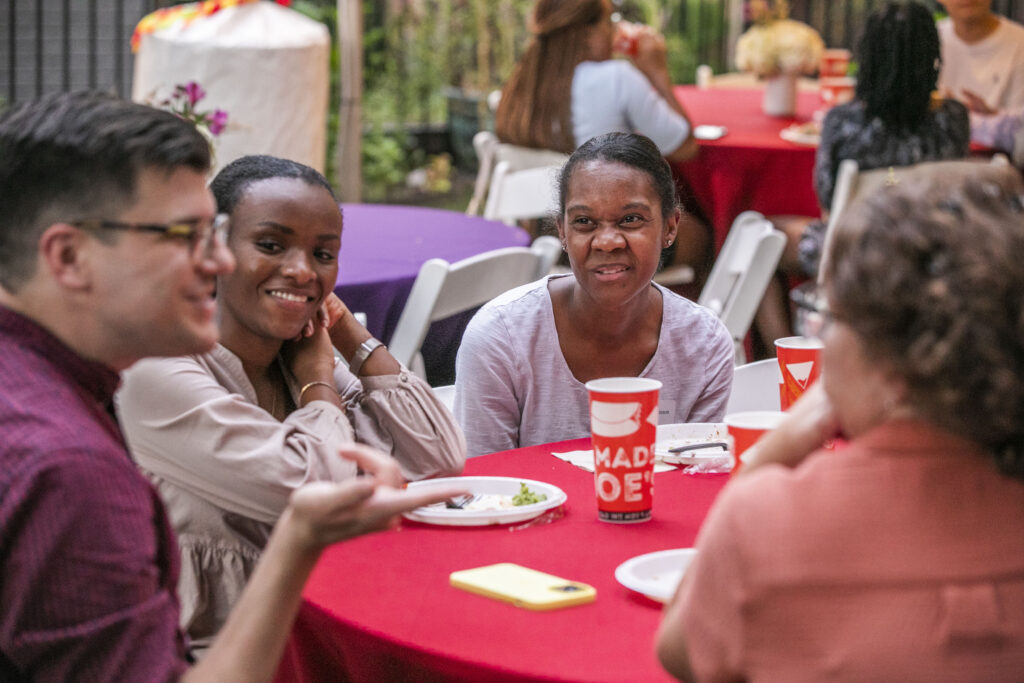 Two Black women listen to a man with glasses speak as they have dinner at a round table