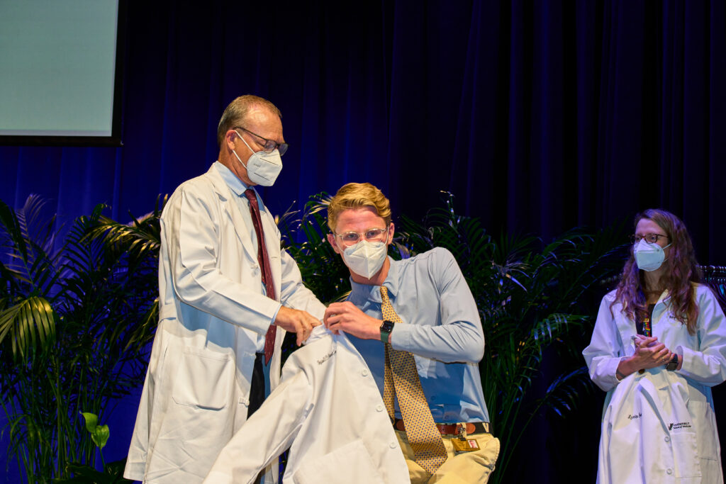 A man in a mask and long white coat helps a younger man put on a short white coat
